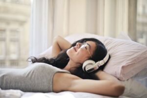 Woman with headphones in a gray tank top lying on a bed.